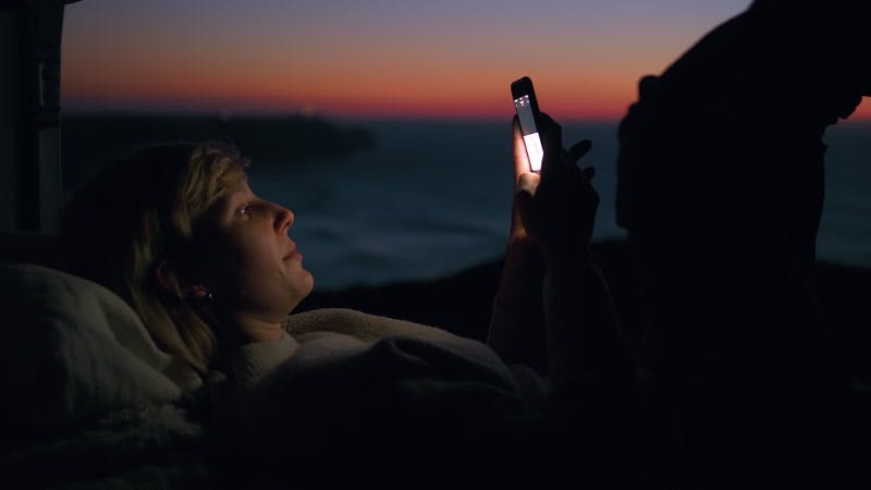 woman lying down near the sea and using a phone at sunset