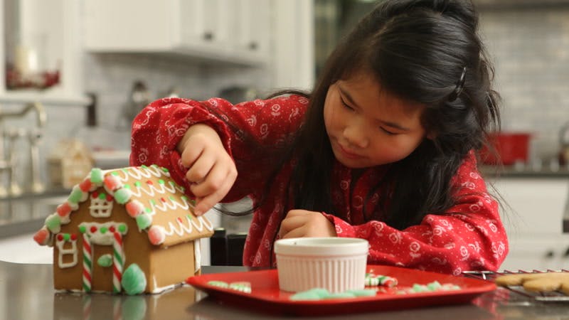 little girl decorating a gingerbread house on a kitchen counter