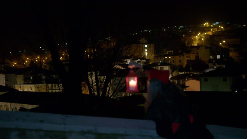 person in an elf costume hanging a lantern from a roof top near town lights at night