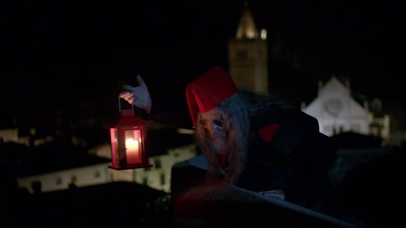 bearded man dressed as an elf and holding a lantern while looking over a rooftop at night
