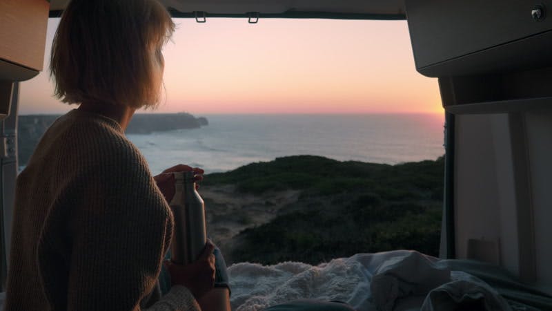 woman sitting in an open camper van and drinking from a flask at sunset