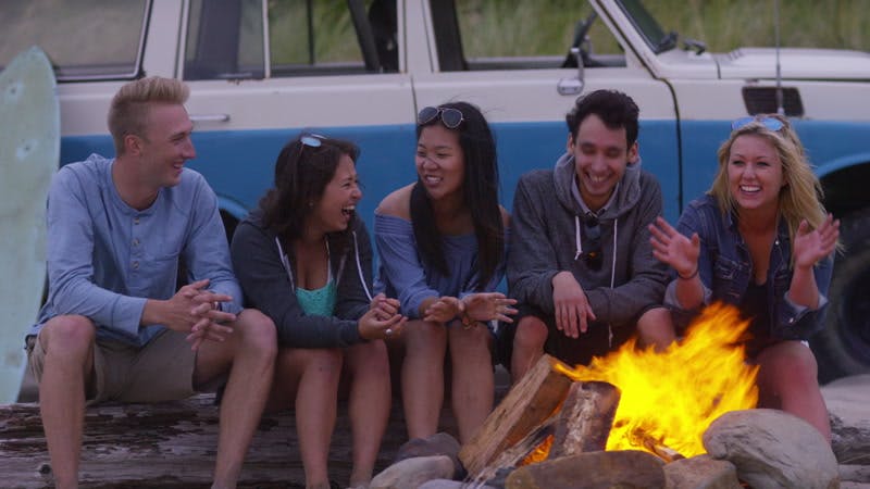 five people sitting by a campfire next to a car on a beach