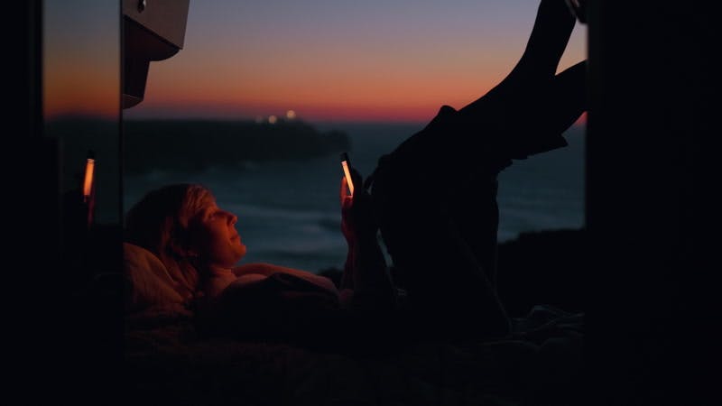 woman lying with legs up and using a phone in an open camper van on the beach at sunset
