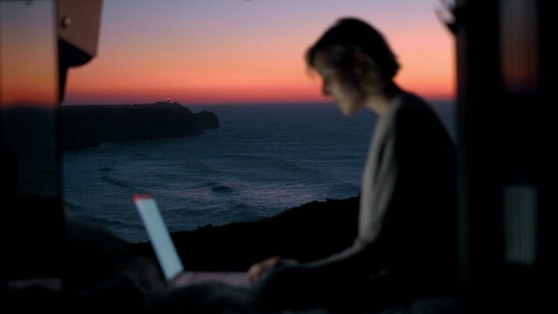 woman typing on a laptop in an open camper van on the coast at sunset