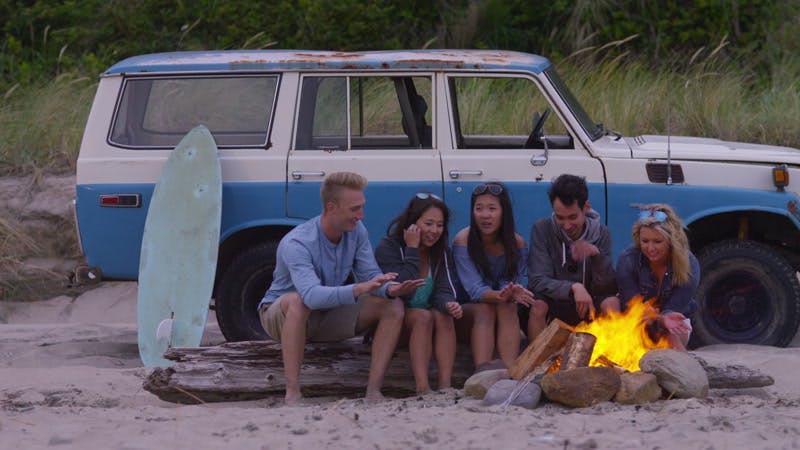 five people sitting by a campfire near a car and a surfboard on a beach